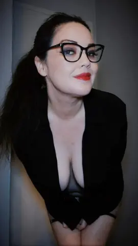 Smiling woman with dark hair and glasses wearing a black blouse in a moody indoor setting.