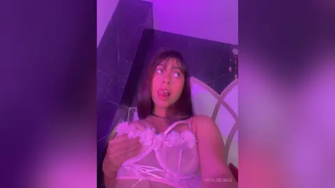 Young woman in white lace lingerie posing in a moody purple lit bedroom setting.