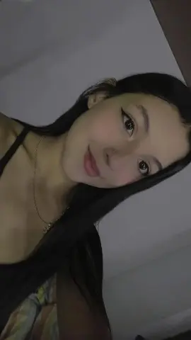 AveryCobb smiles at the camera with long black hair, wearing a black tank top and a gold necklace against a plain white wall.