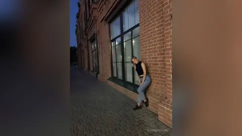 Stylish woman in a black tank top and jeans leaning against a brick building on a city sidewalk at night.