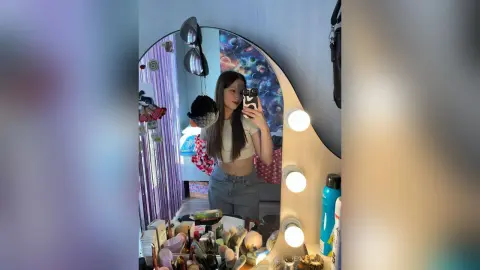 Young woman in a white crop top taking a selfie in a cozy bedroom with a lighted vanity mirror.