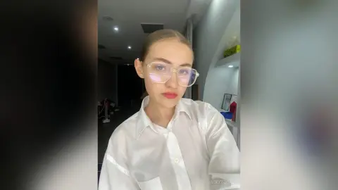 VickyToure wearing clear glasses and a white button-down shirt, looking directly at the camera with her hair pulled back.