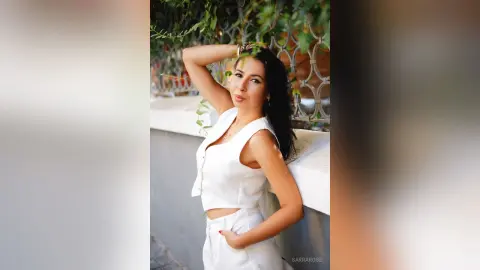 Smiling woman in a white sleeveless top posing outdoors against a lush green garden background.
