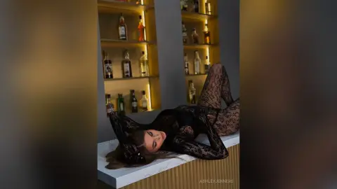 Glamorous woman in a black lace bodysuit lounging on a marble bar in a moody, dimly lit lounge setting.