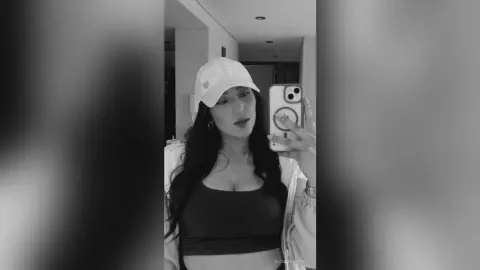 Young woman in a white cap and dark tank top taking a moody black and white mirror selfie indoors.