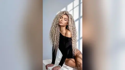 Elegant woman with long wavy blonde hair wearing a black long-sleeve bodysuit in a bright minimalist studio.