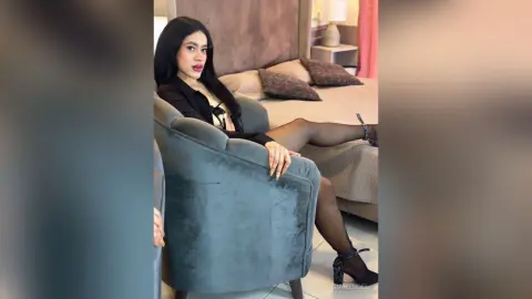 Elegant woman in a black outfit and sheer tights sitting on a grey armchair in a cozy bedroom setting.