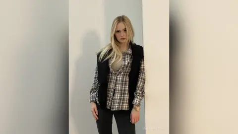 Blonde woman in a plaid shirt and black vest standing against a minimalist white wall with a neutral mood.