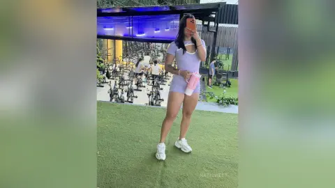 NatiSweet poses in a light purple cutout bodysuit and white sneakers while standing on green turf in a gym setting.