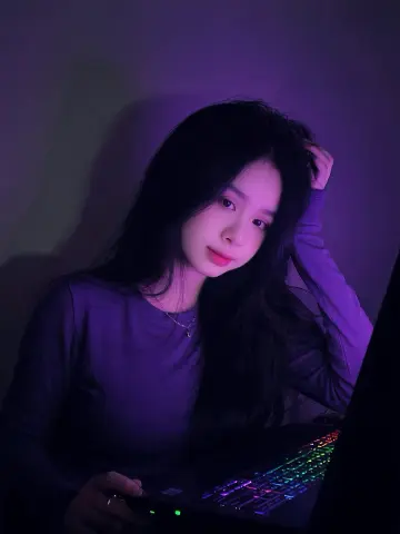 Young woman in a purple long-sleeve shirt working on a laptop in moody, dim violet lighting.