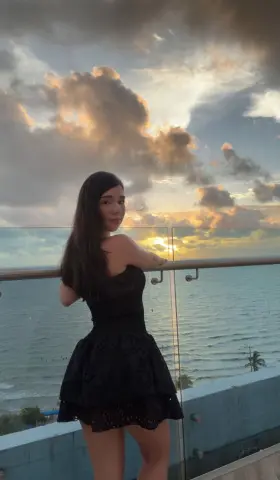 Young woman in a black mini dress standing on a balcony overlooking the ocean during a golden sunset.
