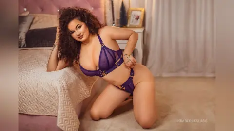 Curvy woman in purple lace lingerie posing on a bed in a bright, cozy bedroom setting.