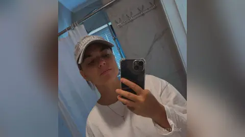 Young woman in a white shirt and grey baseball cap taking a casual mirror selfie in a bright bathroom.