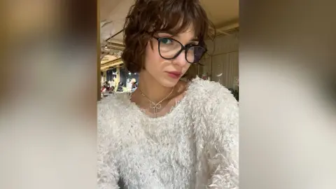 Young woman with short wavy hair and black glasses wearing a cozy white textured sweater in a bright indoor setting.