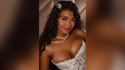 Glamorous woman with wavy dark hair in a white lace outfit posing with a radiant, glowing makeup look.