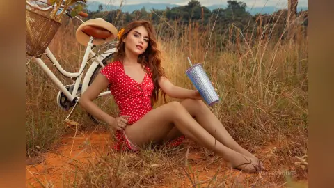 ValeryRouge sits in a grassy field wearing a red polka-dot bodysuit next to a white bicycle and a blue cup.