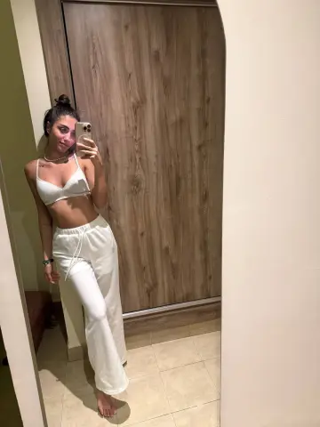 Young woman in white linen crop top and lounge pants taking a mirror selfie in a modern room.