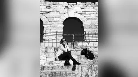 MontseFerrera sits on stone steps wearing a light-colored top, dark pants, and black boots while looking at the camera.