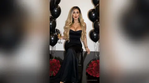 Elegant woman in a black evening gown holding champagne near black balloons and red roses at a glamorous party.