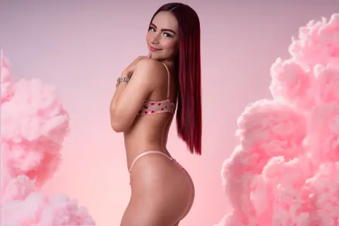 Smiling woman with long red hair in pink lingerie posing amidst dreamy pink clouds.