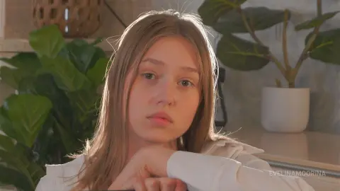 Melancholic young woman with long blonde hair wearing a white shirt in a bright room with green houseplants.
