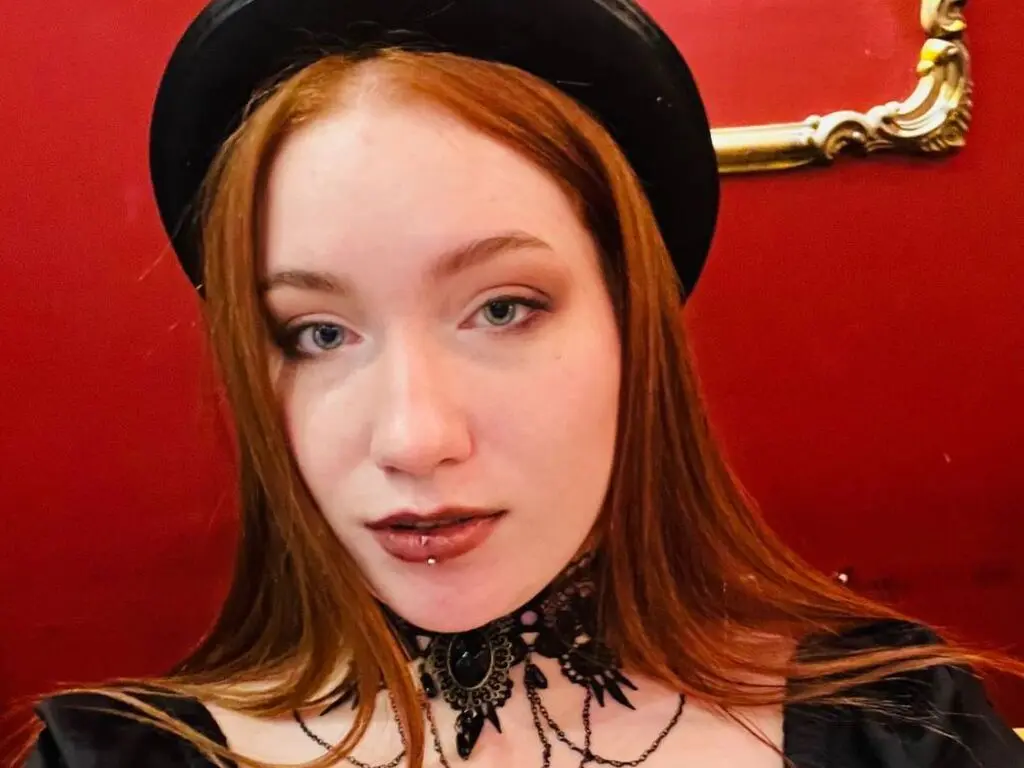 Redheaded woman in gothic black outfit and hat posing against a red wall with vintage gold frame decor.