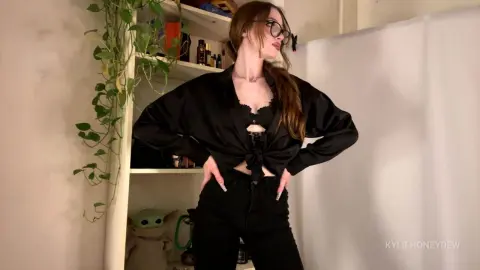 KylieHoneydew poses in a black satin shirt and black jeans, looking away with her hands on her hips in a room with plants.