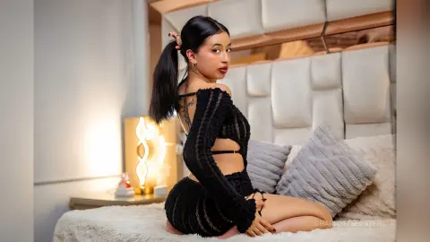 Young woman in a black knit cutout outfit sitting on a bed in a cozy, modern bedroom setting.