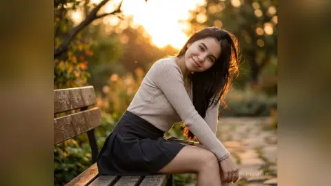 Smiling young woman in a beige sweater and dark skirt sitting on a park bench during a golden hour sunset.
