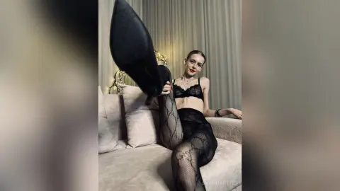 Young woman in black lace lingerie and patterned tights relaxing on a beige sofa in a cozy room.