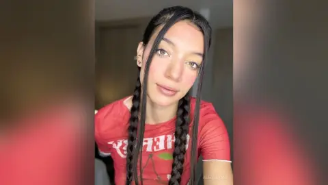 AleDilucas looks at the camera with her black hair in two long braids, wearing a red t-shirt in a close-up shot.