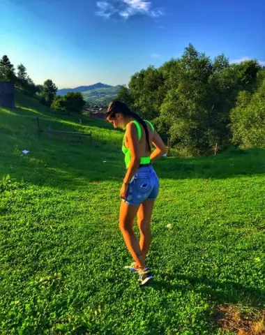 Young woman in a lime green tank top and denim shorts standing in a sunny green meadow under a blue sky.