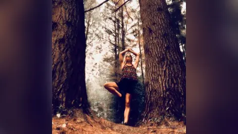 Woman in patterned tank top practicing yoga pose in a misty, sunlit forest setting.