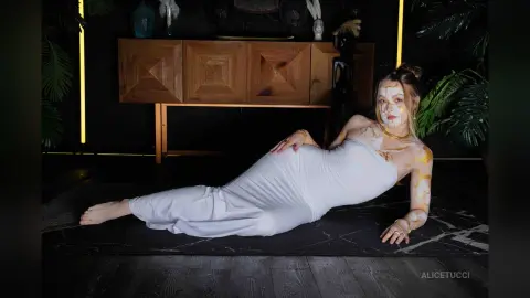 AliceTucci lies on a dark rug wearing a white tube top and white pants, with gold paint or markings visible on her arms, chest, and face.