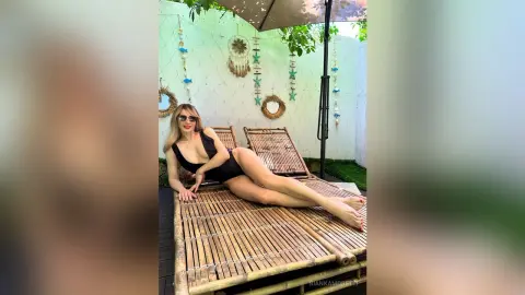 BiankaMoretti wearing a black one-piece swimsuit, lying on a bamboo lounge chair outdoors with sunglasses and blonde hair.