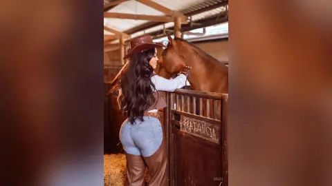 Cowgirl in a brown hat and denim jeans bonding with a brown horse in a rustic stable setting.