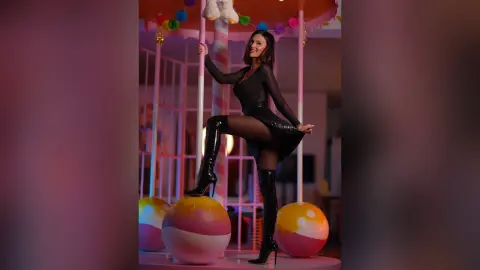 Smiling woman in a black long-sleeve mini dress and tall boots posing in a whimsical pink pastel carnival setting.