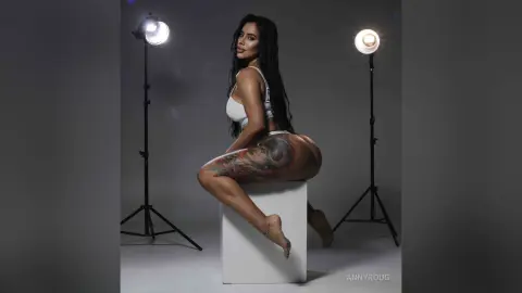 Curvy tattooed woman in white lingerie sitting on a white pedestal in a professional studio setting.