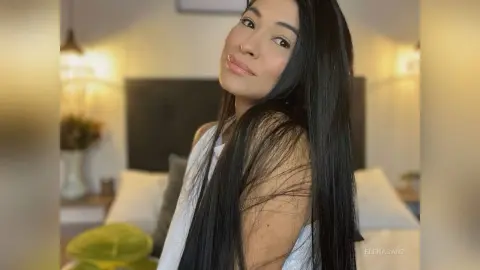 Smiling woman with long black hair wearing a white sleeveless top in a cozy, sunlit bedroom setting.