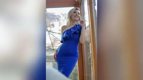 Elegant woman in a royal blue off-the-shoulder cocktail dress posing by a bright window in a dreamy mood.