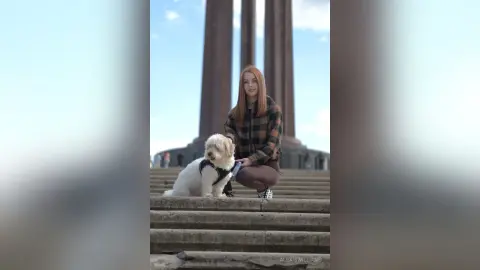 Redheaded woman in a plaid shirt sitting on stone steps with a small white dog in front of tall columns.