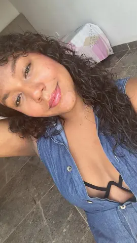 Smiling woman with curly hair wearing a blue denim sleeveless top in a bright indoor setting.