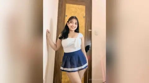 Smiling young woman in a white halter top and navy pleated mini skirt posing in a bright indoor setting.