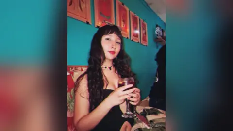 Young woman with long wavy hair in a black dress holding a glass of red wine in a cozy room.
