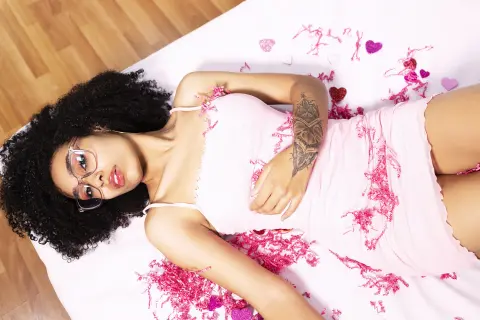 Young woman in a pink slip dress lying on white bedding surrounded by pink heart confetti.