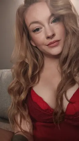 Smiling blonde woman in a red lace top posing in a cozy indoor setting with soft lighting.