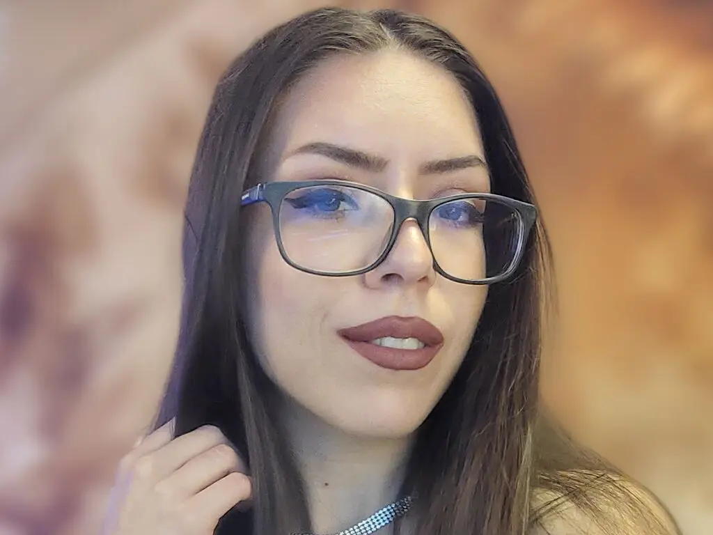 Portrait of a brunette woman wearing black glasses and dark lipstick in a warm, soft-lit indoor setting.