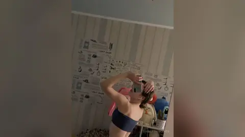 Young woman in a black tube top posing in front of patterned wallpaper for a casual indoor mirror selfie.