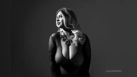 Elegant woman in a sheer black bodysuit and statement necklace posing against a dark studio background.