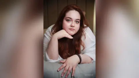 Redheaded woman in a white blouse leaning on a pillow with a calm, serene expression in a soft-lit room.